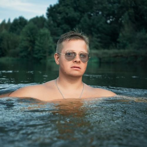 Man with sunglasses enjoys a refreshing swim in a river surrounded by lush greenery.