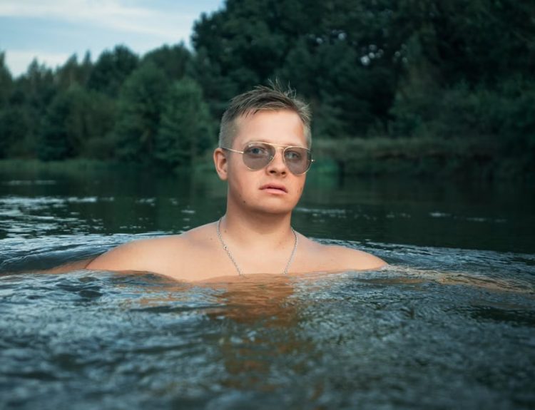 Man with sunglasses enjoys a refreshing swim in a river surrounded by lush greenery.