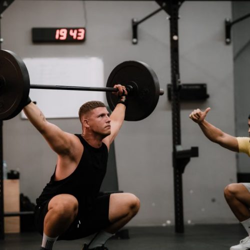 A muscular man performing a weightlifting squat with a coach guiding him indoors.