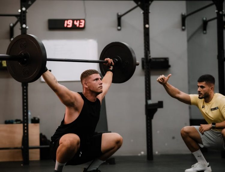 A muscular man performing a weightlifting squat with a coach guiding him indoors.
